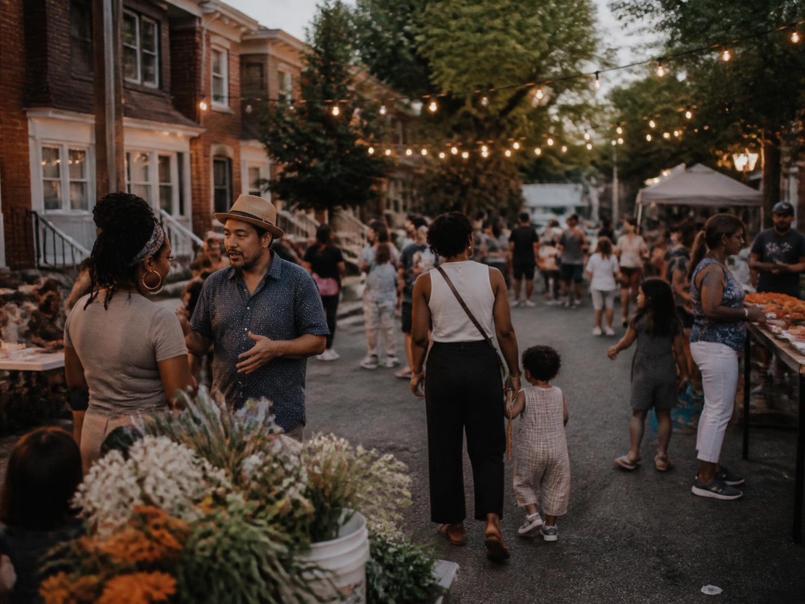 Residents of all ages moving through an evening neighborhood block gathering under string lights