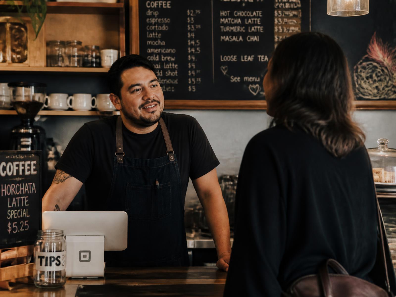 A neighborhood coffee shop owner talking with a regular customer at the counter