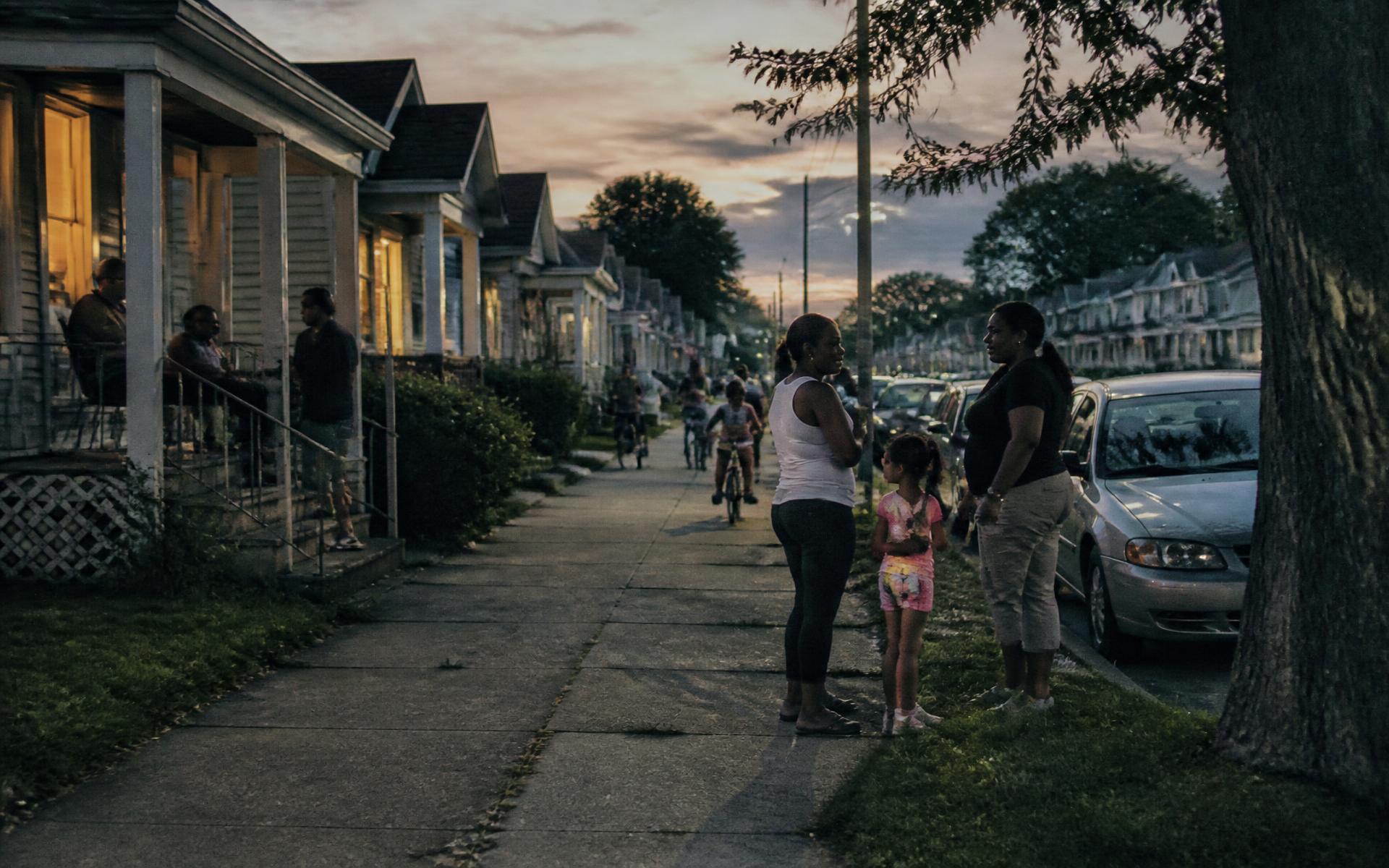 A neighborhood street at dusk with residents on porches and sidewalks and children riding bikes