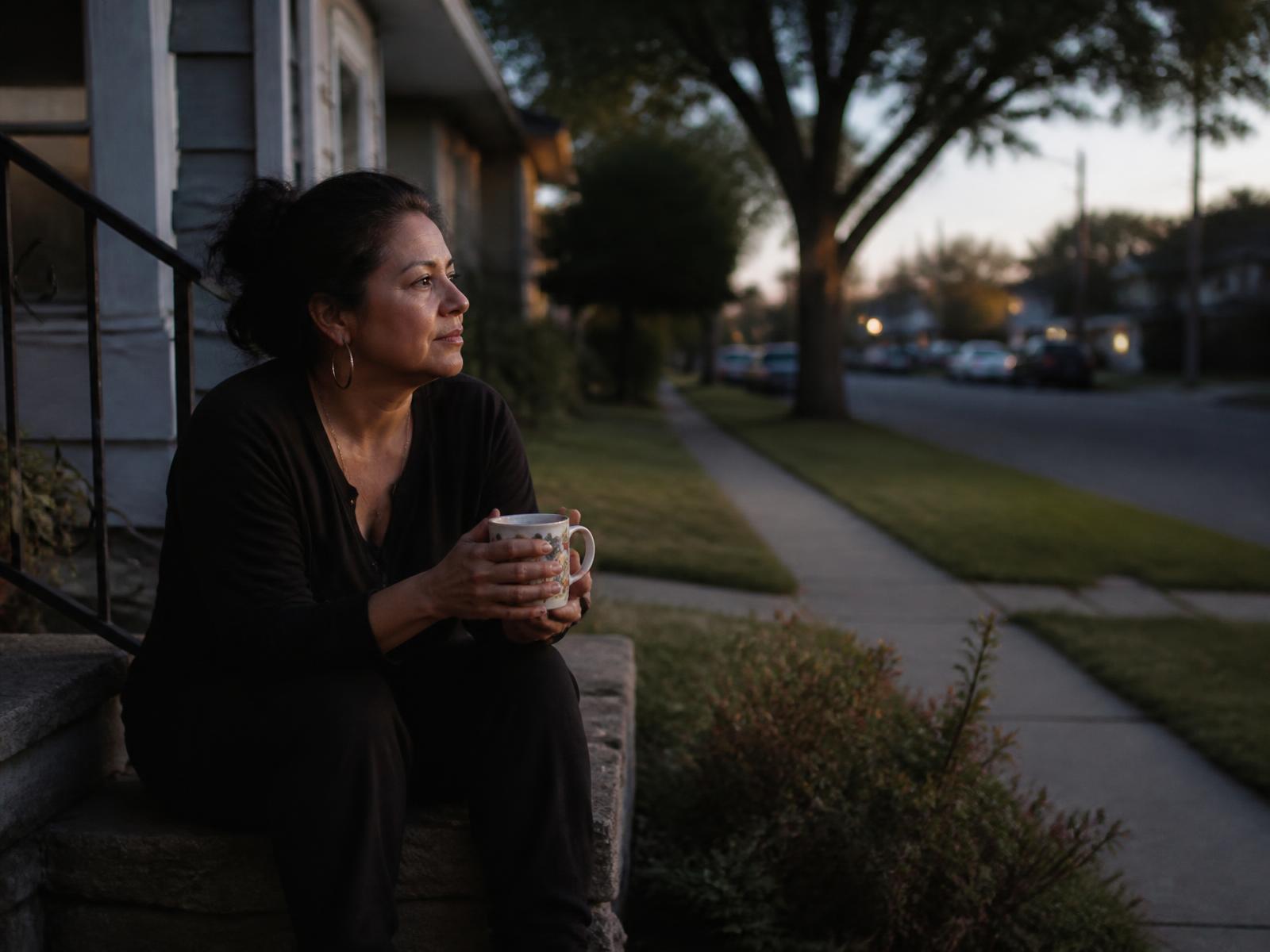 A woman sitting alone on her front porch steps at dusk holding a coffee mug, looking out at the street