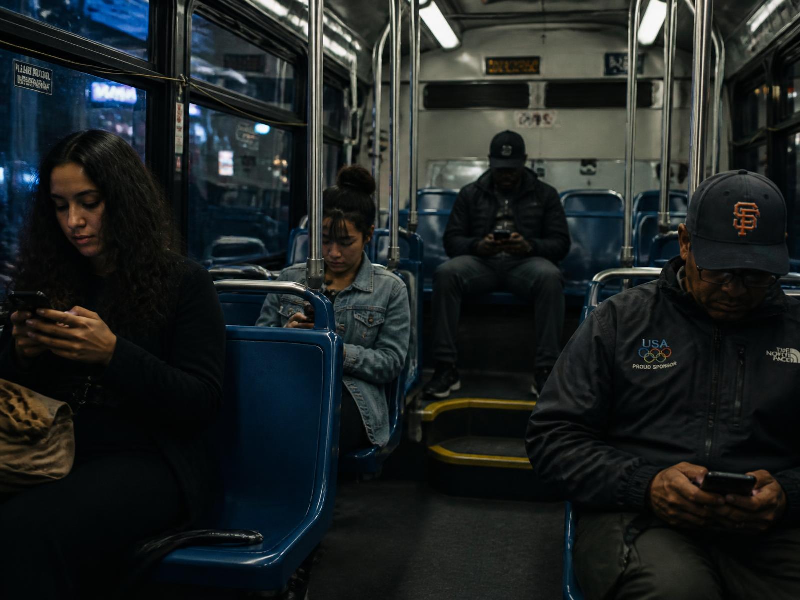 Passengers sitting apart on a city bus at night, each looking at their own phone